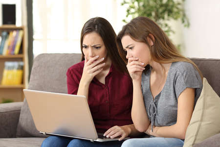 Two worried friends watching media content on line with a pc sitting on a sofa in the living room in a house indoorの写真素材