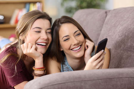 Front view portrait of two happy friends watching media content on line in a smart phone lying on a couch in the living room at homeの写真素材