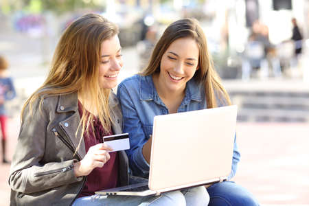 Two friends buying on line with credit card and a laptop sitting in a bench in the street with an urban backgroundの写真素材