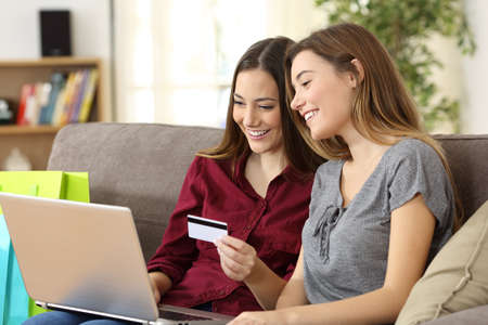 Two happy roommates buying together on line with credit card and a laptop sitting on a sofa at home with a homey backgroundの写真素材
