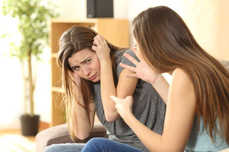 Girl scolding to her scared sister or friend sitting on a couch in the living room in a house interiorの写真素材