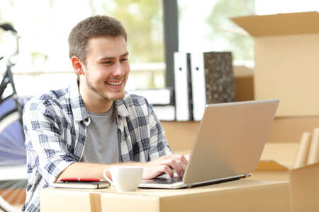 Happy entrepreneur sitting on the floor working on line in his new workplace using a carton box like a desktop while is moving officeの写真素材