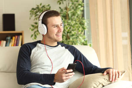 Handsome guy listening music and relaxing sitting on a sofa and looking through the window of the living room at homeの写真素材