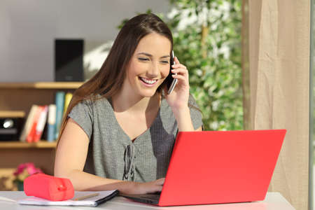 Happy student asking information by phone consulting a laptop on line sitting in a table at home with a colorful backgroundの写真素材