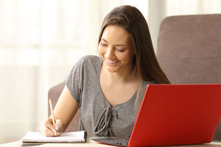 Student learning and taking notes in a paper notebook with a red laptop beside sitting on the floor in the living room at homeの写真素材