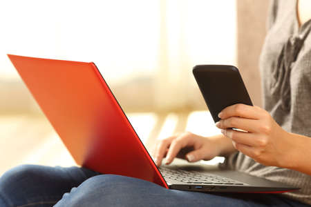 Close up of a girl hands using a phone and a red laptop sitting on the floor of the living room at home with a warm light in the backgroundの写真素材