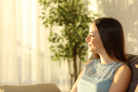 Side view of one woman thinking at sunset sitting on a sofa in the living room in a house interior with a window and a warm light in the backgroundの写真素材
