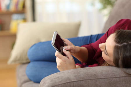 Close up of a relaxed girl using a smart phone lying on a sofa in the living room at home with a cozy backgroundの写真素材