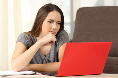 Pensive student studying on line with a red laptop sitting on the floor in the living room at homeの写真素材