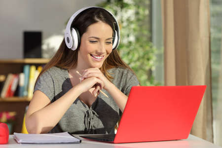 Portrait of a beautiful student viewing and listening video tutorials on line with headphones and a red pc sitting in a table at homeの写真素材