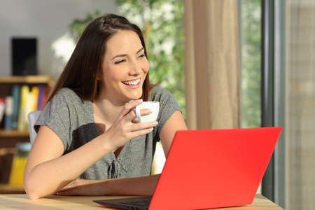 Happy woman with a laptop holding a cup of coffee relaxing and thinking looking through a window at homeの写真素材