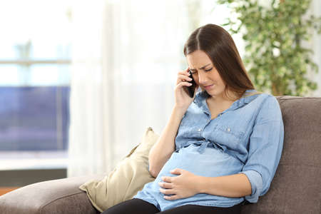 Young pregnant woman suffering belly ache and calling on the phone sitting on a couch in the living room in a house interiorの写真素材