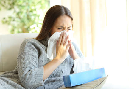 Woman blowing in a wipe suffering flu symptoms sitting on a sofa at home in winterの写真素材