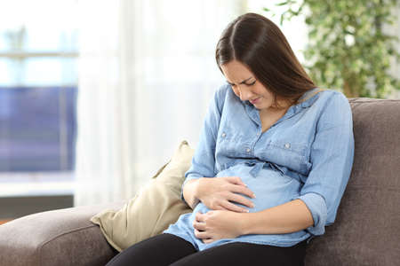 Pregnant woman suffering belly ache sitting on a couch in the living room at homeの写真素材