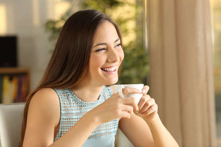 Happy attractive and thoughtful girl holding a mug of coffee looking outdoors through the window with a warm light of sunset at homeの写真素材