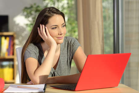 Student thinking and looking away through a window with a red laptop sitting in a table at home with a homey backgroundの写真素材