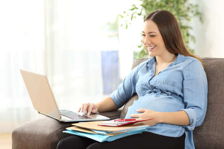 Pregnant entrepreneur working on line with a laptop sitting on a sofa in the living room at homeの写真素材