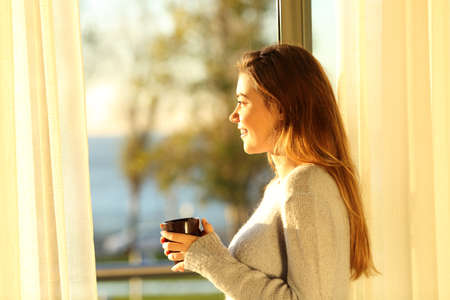 Side view portrait of a single relaxed girl looking the sea outside through a window and holding a...