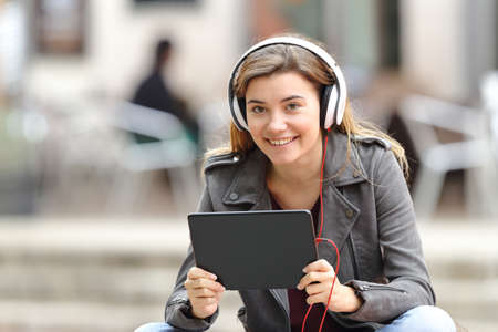 Beautiful girl listening music with headphones and tablet looking at you sitting in a bench in the streetの写真素材