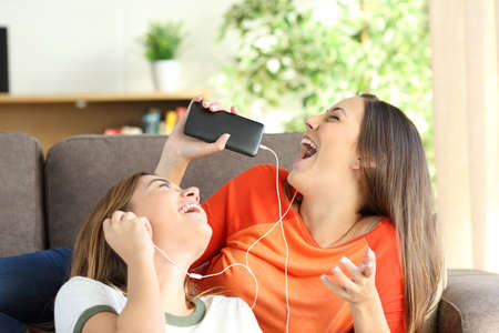 Excited friends or sisters listening music and singing on a couch in the living room at homeの写真素材