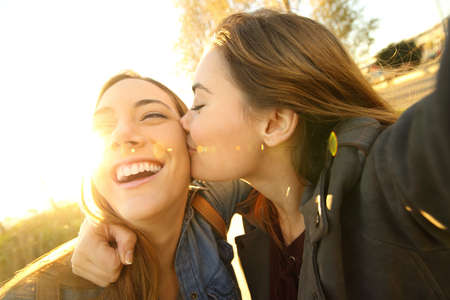 Two affectionate friends kissing and taking a selfie outdoors in the street at sunset with a warm light in the backgroundの写真素材