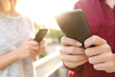 Two girls using everyone her smart phone texting messages outdoors at sunset with a park in the backgroundの写真素材