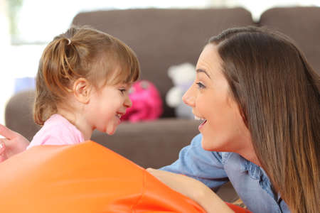 Side view portrait of a happy mother and 2 years baby daughter facing and joking in the living room at homeの写真素材