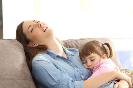 Tired mother sleeping embracing to her asleep baby daughter sitting on a sofa at homeの写真素材