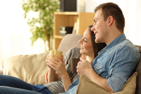 Couple looking away through the window sitting on a comfortable sofa in a homey living room with a warm light in the backgroundの写真素材
