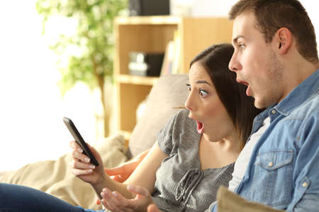 Astonished couple checking amazing content on line in a smart phone sitting on a couch in the living room in a house interiorの写真素材