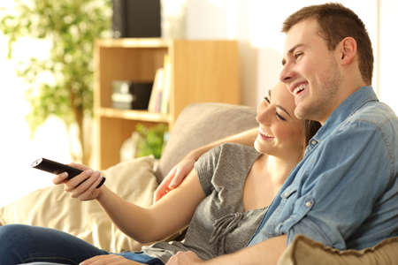 Happy couple watching tv and holding a remote control sitting on a sofa in the living room at homeの写真素材
