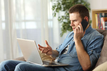 Angry man having problems on line with a laptop talking on the mobile phone with support service sitting on a couch in the living room at home