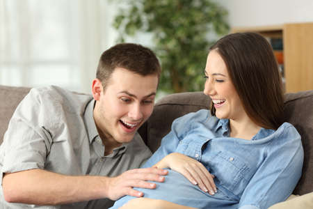 Happy husband and pregnant woman expecting birth sitting on a couch in the living room at homeの写真素材
