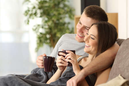 Happy couple relaxing and drinking tea covered with a blanket sitting on a sofa in the living room in a cozy house interiorの写真素材