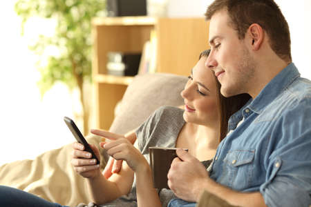 Serious couple checking content in a smart phone sitting on a sofa in the living room at home with a warm lightの写真素材
