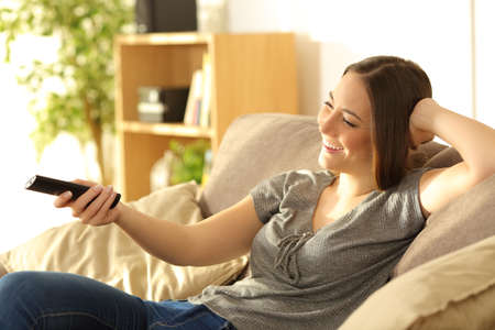 Happy girl watching tv and holding the remote control sitting on a sofa in the living room at homeの写真素材