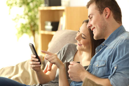 Happy couple checking a smart phone sitting on a couch in the living room at homeの写真素材