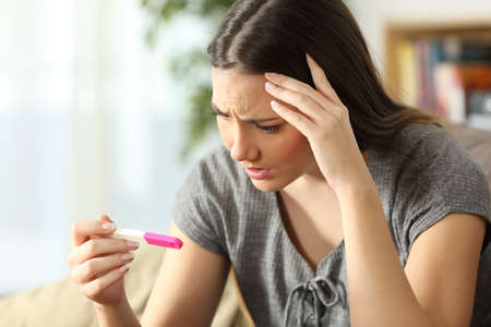 Worried woman checking a pregnancy test sitting on a couch in the living room at homeの写真素材