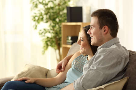 Side view of a happy couple relaxing and looking through a window sitting on a sofa in the living room at homeの写真素材