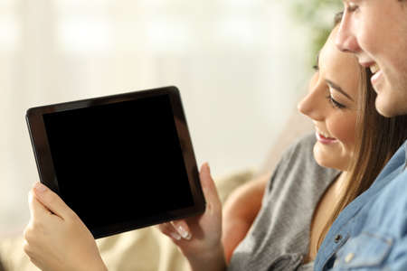 Close up of a happy couple watching media in a tablet and showing the blank screen sitting on a sofa in the living room at home