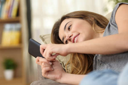 Relaxed teen texting in a smart phone lying on sofa in the living room at homeの写真素材