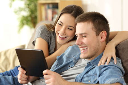 Couple laughing watching streaming content in a tablet sitting on a sofa in the living room at home with a warm lightの写真素材