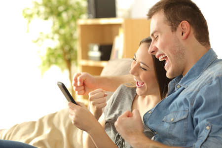 Excited couple checking good news in a smart phone sitting on a sofa in the living room at homeの写真素材
