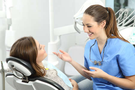Dentist wearing blue coat explaining procedure to a patient who is sitting on a chair in a clinic box before treatmentの写真素材