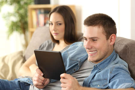 Husband watching online content in a tablet and ignoring to his angry wife who is seated beside him on a sofa at homeの写真素材