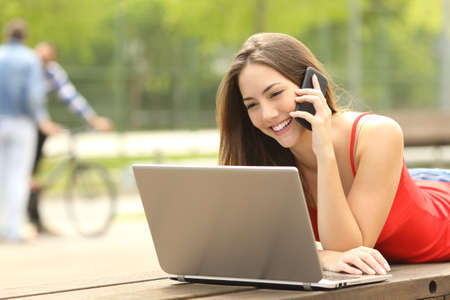 Single student on line with a laptop and calling customer service on the phone lying on a bench in the street with a green backgroundの写真素材