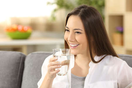 Single woman drinking water from a glass sitting on a sofa in the living room at homeの写真素材