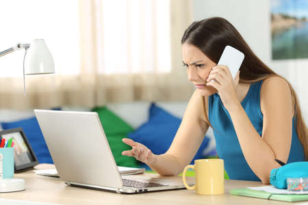 Angry student looking at a laptop and consulting on phone with customer service in her bedroom in a house interiorの写真素材