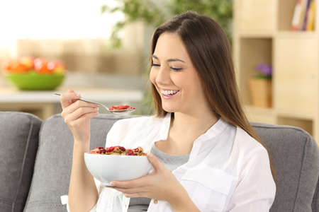 Cheerful woman eating cereals sitting on a sofa in the living room at homeの写真素材