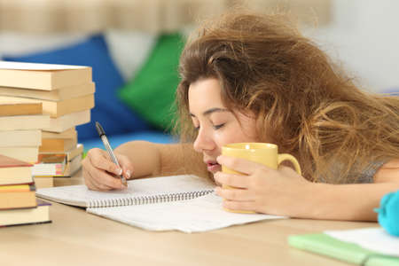 Tired and sleepy student with tousled hair trying to write notes on a desk in her room in a house indoorの写真素材